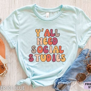 May include: A light blue t-shirt with the text "Y'all need social studies" in colorful retro-style lettering.