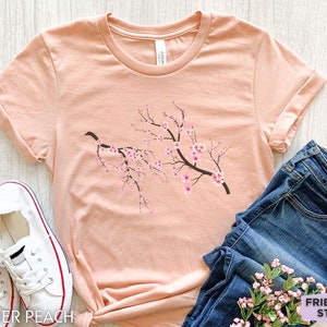 Puede incluir: Una camiseta de color melocot&oacute;n con un gr&aacute;fico de una rama de flores de cerezo rosas. La camiseta est&aacute; colocada sobre una superficie de madera clara con un par de zapatillas blancas y unos vaqueros azules.