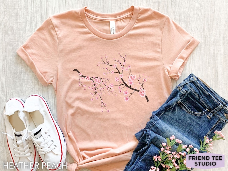 Puede incluir: Una camiseta de color melocot&oacute;n con un gr&aacute;fico de una rama de flores de cerezo rosas. La camiseta est&aacute; colocada sobre una superficie de madera clara con un par de zapatillas blancas y unos vaqueros azules.