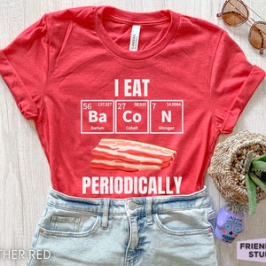 Puede incluir: Camiseta rojo jaspeado con el texto blanco "I EAT BACON PERIODICALLY". El dise&ntilde;o presenta una tira de tocino y los elementos Ba, Co y N de la tabla peri&oacute;dica. La camiseta es de Friend Tee Studio.