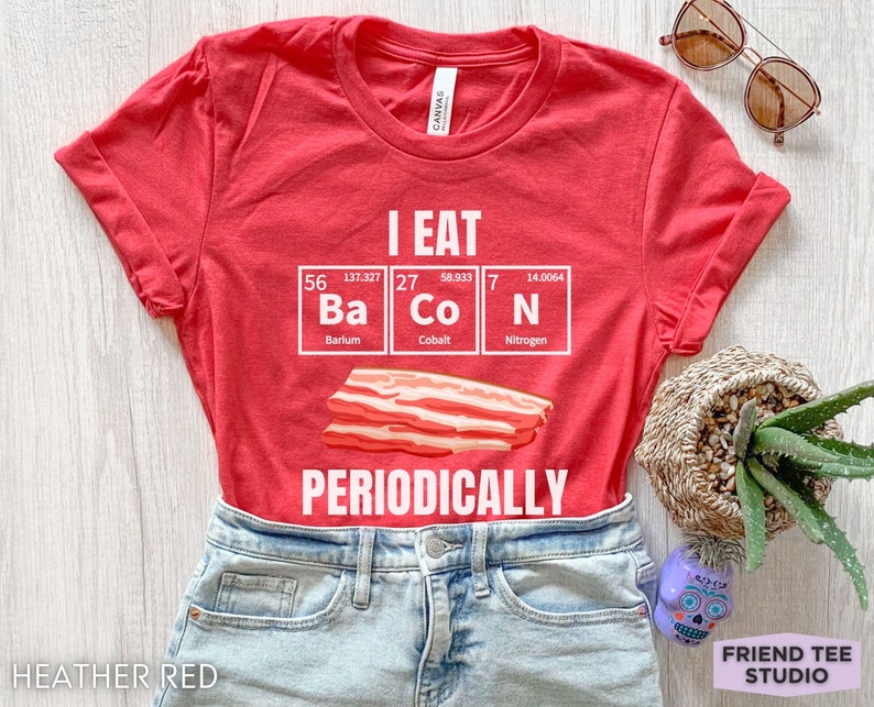 Puede incluir: Camiseta rojo jaspeado con el texto blanco "I EAT BACON PERIODICALLY". El dise&ntilde;o presenta una tira de tocino y los elementos Ba, Co y N de la tabla peri&oacute;dica. La camiseta es de Friend Tee Studio.