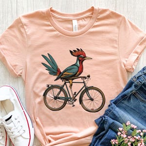 K&ouml;nnte beinhalten: Ein pfirsichfarbenes T-Shirt mit einem bunten Vogel, der Fahrrad f&auml;hrt. Der Vogel hat einen roten Kamm, blaue und gr&uuml;ne Federn und sitzt auf einem schwarzen Fahrrad mit braunen Reifen. Das Shirt wird mit blauen Jeans und wei&szlig;en Turnschuhen kombiniert.
