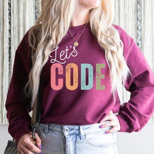 K&ouml;nnte beinhalten: Burgunderfarbenes Sweatshirt mit dem Text "Let's CODE" in Wei&szlig;, Rosa, Orange, T&uuml;rkis und Gr&uuml;n. Das Sweatshirt ist ein lang&auml;rmeliger Rundhalsausschnitt. Die Person tr&auml;gt eine blaue Jeans und eine goldene Halskette.