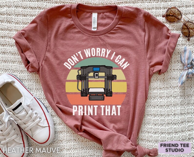 May include: A heather mauve short-sleeve t-shirt featuring a graphic of a 3D printer centered over a retro-style sunset circle in shades of green, yellow, and orange. The white text above and below the printer reads, "DON'T WORRY I CAN PRINT THAT."