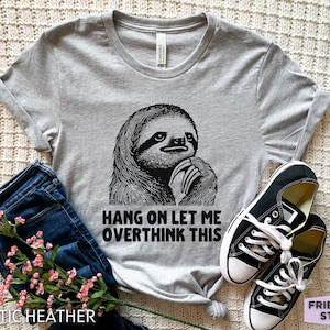May include: A light grey short-sleeved t-shirt featuring a black graphic illustration of a sloth resting its chin on its hand. Below the sloth, the text reads, HANG ON LET ME OVERTHINK THIS. The shirt is paired with dark blue denim jeans and black trainers.
