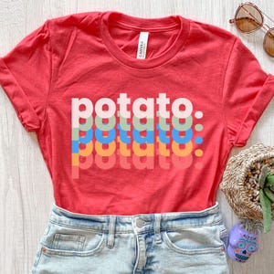 May include: A red short-sleeve t-shirt featuring the word potato printed in five stacked, overlapping rows of different colors including white, light green, blue, orange, and pink. The shirt has a crew neckline and a white interior brand label.