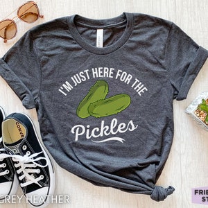 May include: Dark grey heather t-shirt with the text "I'M JUST HERE FOR THE PICKLES" and a graphic of two green pickles. The shirt is styled with sunglasses and black sneakers.
