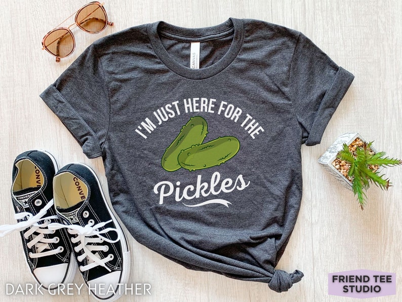 May include: Dark grey heather t-shirt with the text "I'M JUST HERE FOR THE PICKLES" and a graphic of two green pickles. The shirt is styled with sunglasses and black sneakers.