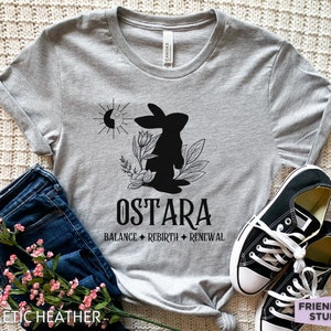Puede incluir: Una camiseta gris jaspeado con un gr&aacute;fico negro de un conejo, flores y un s&iacute;mbolo de sol y luna. El texto dice OSTARA, BALANCE, REBIRTH y RENEWAL. La camiseta se combina con pantalones vaqueros azul oscuro y zapatillas de lona negras y blancas.