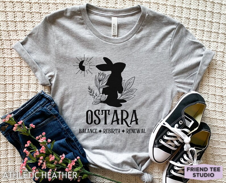Puede incluir: Una camiseta gris jaspeado con un gr&aacute;fico negro de un conejo, flores y un s&iacute;mbolo de sol y luna. El texto dice OSTARA, BALANCE, REBIRTH y RENEWAL. La camiseta se combina con pantalones vaqueros azul oscuro y zapatillas de lona negras y blancas.