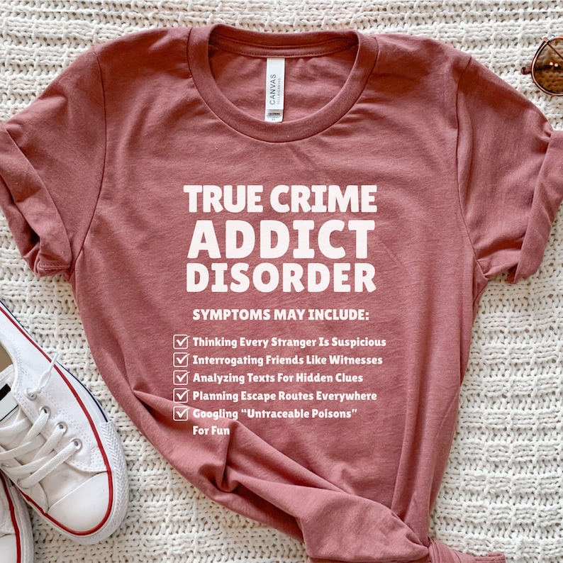 May include: A rose-coloured t-shirt with the text "TRUE CRIME ADDICT DISORDER" in white. The shirt lists symptoms including suspecting strangers, interrogating friends, analysing texts, planning escape routes, and Googling poisons.