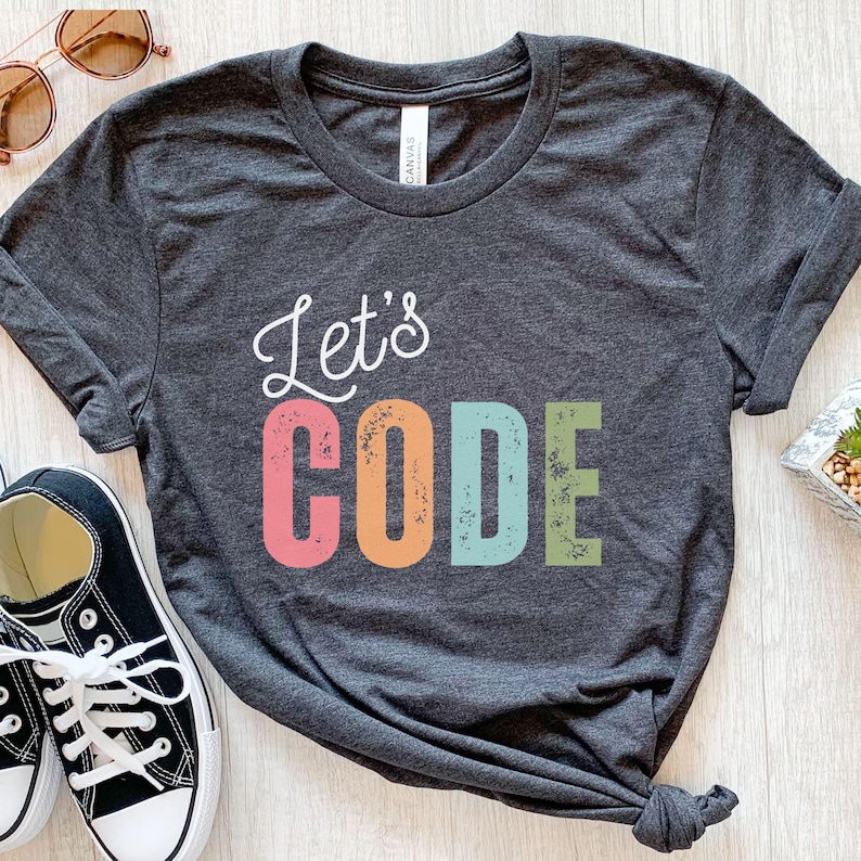 K&ouml;nnte beinhalten: Dunkelgraues T-Shirt mit dem Text "Let's CODE" in Wei&szlig;, Rosa, Orange, Blau und Gr&uuml;n. Das Shirt ist mit schwarz-wei&szlig;en Turnschuhen und Sonnenbrillen versehen.