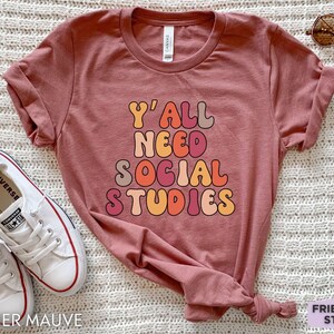 May include: Heather mauve t-shirt with the text "Y'all need social studies" in a retro font with colorful letters.