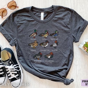 Peut inclure: Un t-shirt gris chin&eacute; fonc&eacute; avec une impression graphique repr&eacute;sentant huit esp&egrave;ces de canards diff&eacute;rentes, accompagn&eacute;es de leurs noms inscrits en dessous : Mallard, Wood, Baikal Teal, Gadwall, Eurasian Teal, Redhead, Rosy-billed Pochard et Red-crested Pochard.