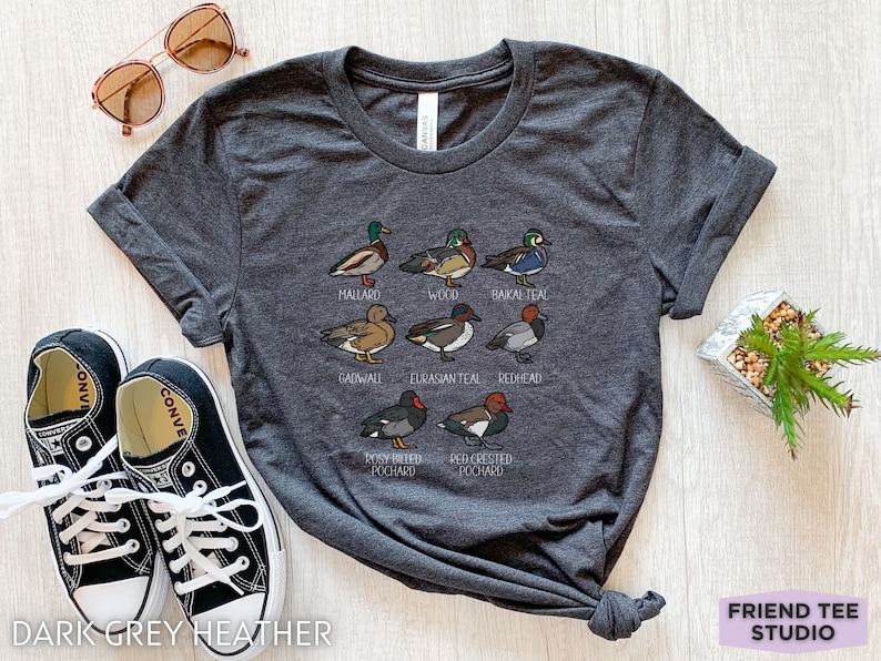Peut inclure: Un t-shirt gris chin&eacute; fonc&eacute; avec une impression graphique repr&eacute;sentant huit esp&egrave;ces de canards diff&eacute;rentes, accompagn&eacute;es de leurs noms inscrits en dessous : Mallard, Wood, Baikal Teal, Gadwall, Eurasian Teal, Redhead, Rosy-billed Pochard et Red-crested Pochard.