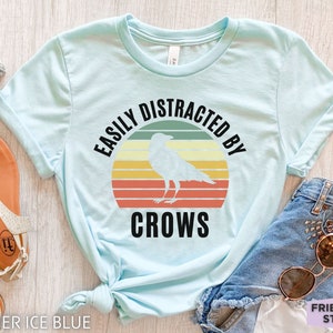 May include: A light blue t-shirt with a retro-style graphic that says "Easily Distracted By Crows" with a black crow silhouette in the center.