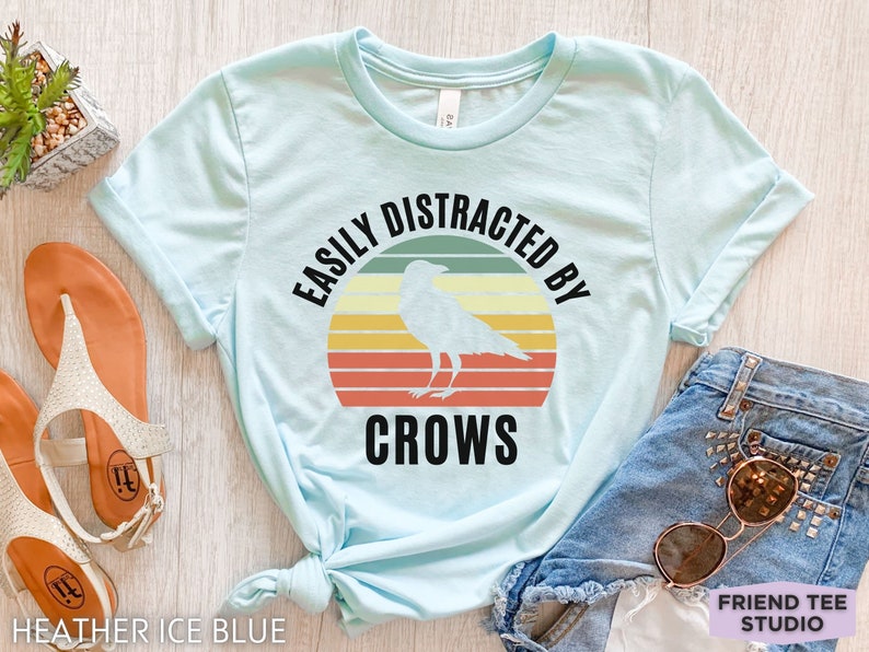 May include: A light blue t-shirt with a retro-style graphic that says "Easily Distracted By Crows" with a black crow silhouette in the center.