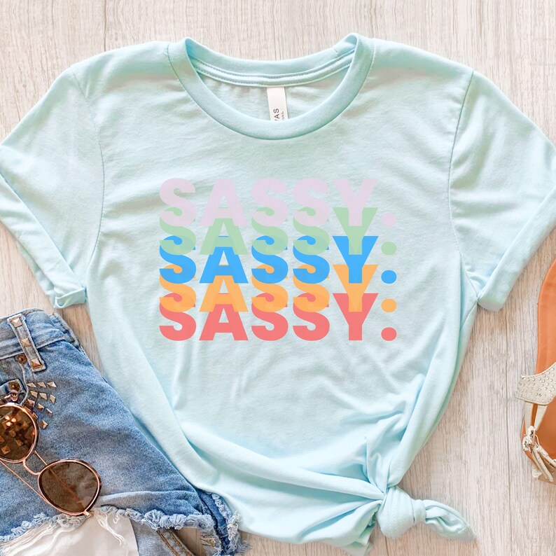 May include: A light blue t-shirt with the word "SASSY" stacked in a colorful, layered design. The word is in pastel colors: purple, green, blue, orange, and red. The shirt is styled with denim shorts and sunglasses.