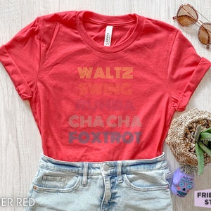 May include: Heather red t-shirt with rolled sleeves. The shirt features the words "Waltz, Swing, Rumba, Cha Cha, Foxtrot" in a retro font. Sunglasses and a small potted plant are visible.