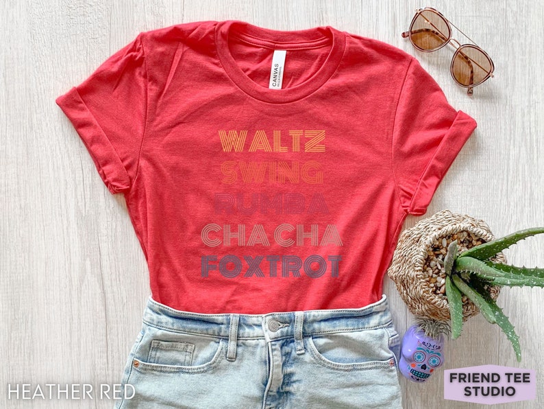 May include: Heather red t-shirt with rolled sleeves. The shirt features the words "Waltz, Swing, Rumba, Cha Cha, Foxtrot" in a retro font. Sunglasses and a small potted plant are visible.