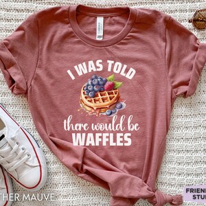 May include: Heather mauve t-shirt with the text "I WAS TOLD there would be WAFFLES" and a waffle graphic topped with blueberries and a raspberry. The shirt is a crew neck and has short sleeves.
