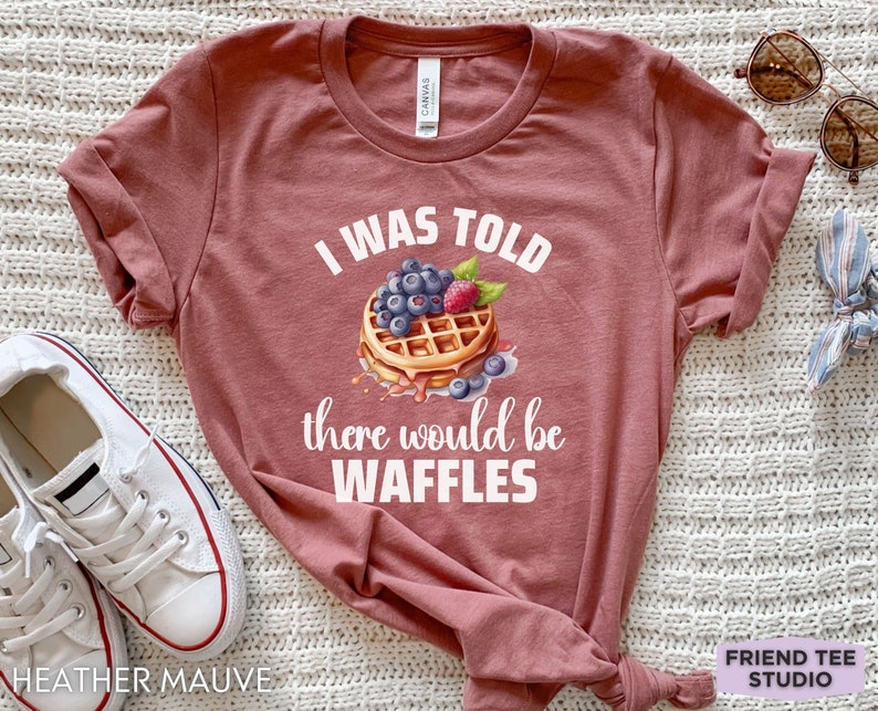 May include: Heather mauve t-shirt with the text "I WAS TOLD there would be WAFFLES" and a waffle graphic topped with blueberries and a raspberry. The shirt is a crew neck and has short sleeves.
