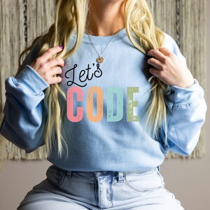 K&ouml;nnte beinhalten: Hellblaues Sweatshirt mit dem Schriftzug "Let's CODE" in einer farbenfrohen, verwitterten Schriftart. Der Text ist in Schwarz, Rosa, Orange, T&uuml;rkis und Gr&uuml;n gehalten. Das Sweatshirt hat lange &Auml;rmel und einen Rundhalsausschnitt.