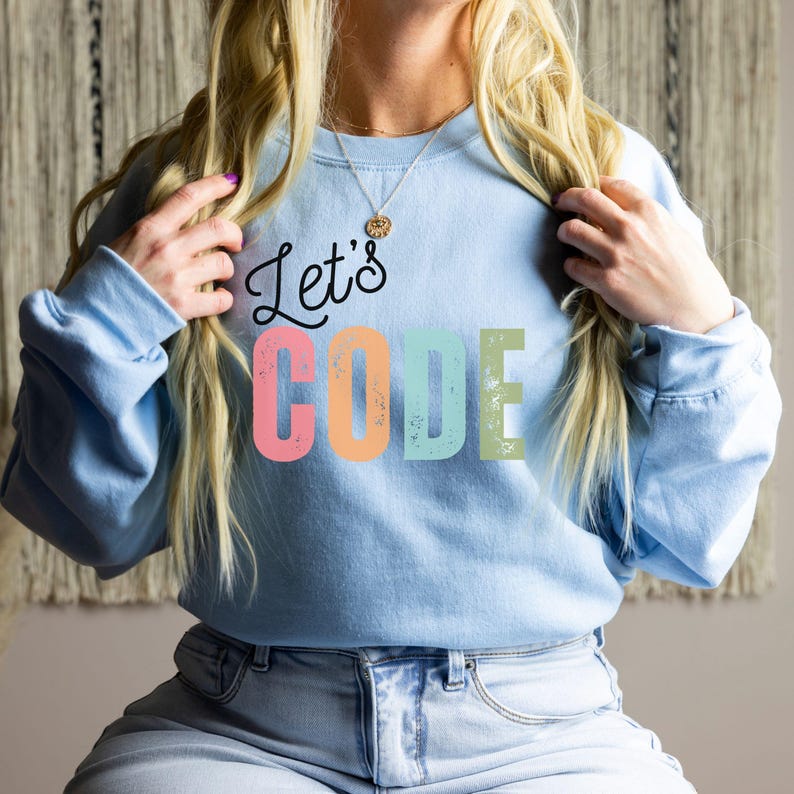K&ouml;nnte beinhalten: Hellblaues Sweatshirt mit dem Schriftzug "Let's CODE" in einer farbenfrohen, verwitterten Schriftart. Der Text ist in Schwarz, Rosa, Orange, T&uuml;rkis und Gr&uuml;n gehalten. Das Sweatshirt hat lange &Auml;rmel und einen Rundhalsausschnitt.