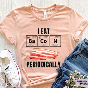 Puede incluir: Camiseta color melocot&oacute;n con el texto negro "I EAT Ba Co N PERIODICALLY" y una ilustraci&oacute;n de bacon. El dise&ntilde;o incorpora los elementos de la tabla peri&oacute;dica Bario, Cobalto y Nitr&oacute;geno. La camiseta se combina con vaqueros azules y zapatillas blancas.