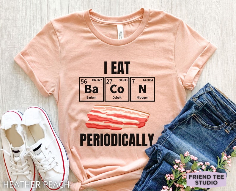 Puede incluir: Camiseta color melocot&oacute;n con el texto negro "I EAT Ba Co N PERIODICALLY" y una ilustraci&oacute;n de bacon. El dise&ntilde;o incorpora los elementos de la tabla peri&oacute;dica Bario, Cobalto y Nitr&oacute;geno. La camiseta se combina con vaqueros azules y zapatillas blancas.