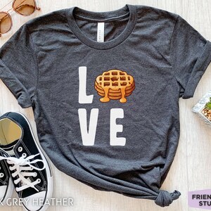 K&ouml;nnte beinhalten: Dunkelgraues T-Shirt mit dem Wort "LOVE" in wei&szlig;. Das "O" ist durch ein Cartoon-Bild eines Waffels mit Sirup ersetzt.