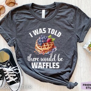 May include: Dark grey heather t-shirt with the text "I WAS TOLD there would be WAFFLES" and a waffle graphic topped with blueberries and raspberries. The shirt also has a pair of black sneakers and sunglasses.