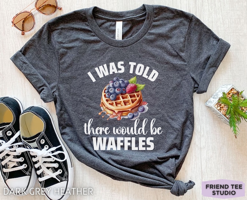 May include: Dark grey heather t-shirt with the text "I WAS TOLD there would be WAFFLES" and a waffle graphic topped with blueberries and raspberries. The shirt also has a pair of black sneakers and sunglasses.