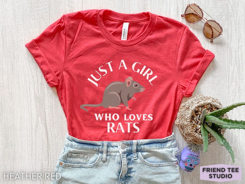 May include: A heather red short-sleeve t-shirt featuring a graphic of a brown rat centered between the white text that reads JUST A GIRL WHO LOVES RATS. The shirt has a crew neckline and is paired with light blue denim jeans with a metal button.