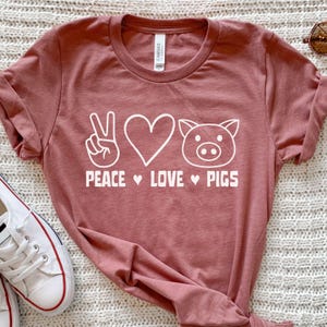 May include: A dusty rose t-shirt with the words "PEACE LOVE PIGS" in white, along with a peace sign, a heart, and a pig face. The shirt is made of soft material and has a crew neck.
