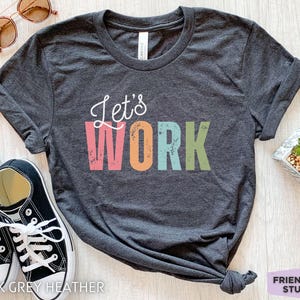 Può includere: Una t-shirt a girocollo grigio scuro mélange con la scritta in corsivo bianco Let's e il testo in stampatello WORK in colori rosa, arancione, azzurro e verde dall'effetto usurato. La t-shirt è presentata con scarpe da ginnastica nere e bianche.