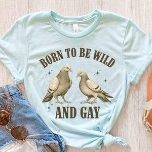May include: Light blue t-shirt with the text "BORN TO BE WILD AND GAY" in brown lettering. The shirt features a graphic of two pigeons. The shirt is tied in a knot at the bottom.