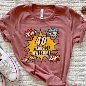 May include: A dusty rose t-shirt featuring a comic book style design for a 40th birthday. The graphic includes the words "POW!", "WOW!", "BOOM!", and "ZAP!" around the text "40 YEARS OF AWESOME".