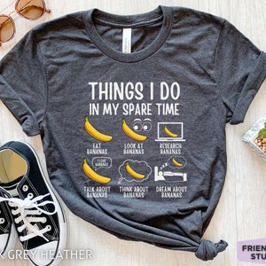 Peut inclure: T-shirt en gris chin&eacute; fonc&eacute; avec un motif blanc qui dit "Things I do in my spare time" et pr&eacute;sente une s&eacute;rie d'illustrations de bananes avec du texte sur le fait de manger, de regarder, de rechercher, de parler, de penser et de r&ecirc;ver de bananes.