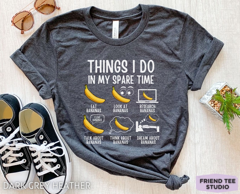 Peut inclure: T-shirt en gris chin&eacute; fonc&eacute; avec un motif blanc qui dit "Things I do in my spare time" et pr&eacute;sente une s&eacute;rie d'illustrations de bananes avec du texte sur le fait de manger, de regarder, de rechercher, de parler, de penser et de r&ecirc;ver de bananes.