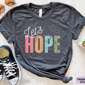 Può includere: Una t-shirt grigio scuro mélange con il testo corsivo bianco Let's e le lettere stampatello HOPE in rosa, arancione, azzurro e verde con una texture usurata. La maglietta è mostrata con scarpe da ginnastica bianche e nere e occhiali da sole.
