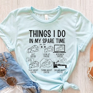 Puede incluir: Una camiseta azul claro con un dise&ntilde;o gr&aacute;fico que dice "Things I do in my spare time" y presenta una serie de actividades relacionadas con el queso, incluyendo comer, mirar, investigar, hablar, pensar y so&ntilde;ar con queso.