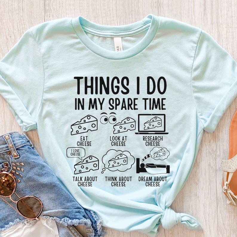 Puede incluir: Una camiseta azul claro con un dise&ntilde;o gr&aacute;fico que dice "Things I do in my spare time" y presenta una serie de actividades relacionadas con el queso, incluyendo comer, mirar, investigar, hablar, pensar y so&ntilde;ar con queso.