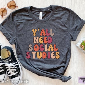 May include: Dark grey heather t-shirt with the text "Y'all need social studies" in colorful retro font.