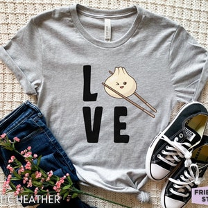 Op de afbeelding: Grijs T-shirt met het woord "LOVE" in zwarte letters. De "O" is vervangen door een cartoon-dumpling met eetstokjes.