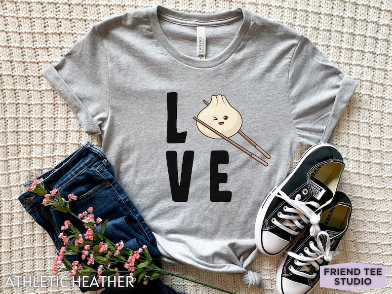 Op de afbeelding: Grijs T-shirt met het woord "LOVE" in zwarte letters. De "O" is vervangen door een cartoon-dumpling met eetstokjes.