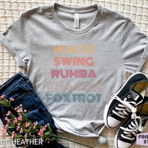 May include: A gray athletic heather t-shirt with the words "Waltz, Swing, Rumba, Cha Cha, Foxtrot" in a retro font. The shirt is paired with blue jeans, black sneakers, and pink flowers.