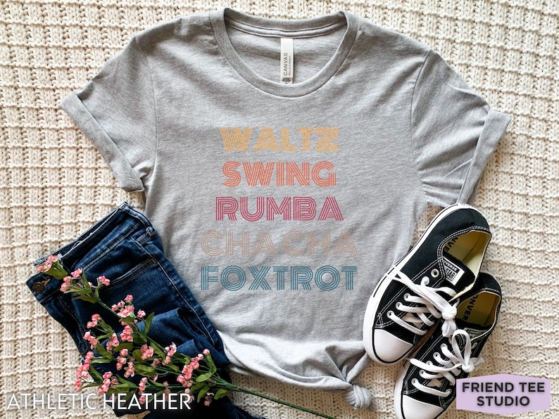 May include: A gray athletic heather t-shirt with the words "Waltz, Swing, Rumba, Cha Cha, Foxtrot" in a retro font. The shirt is paired with blue jeans, black sneakers, and pink flowers.