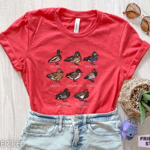 Peut inclure: Un t-shirt &agrave; manches courtes rouge chin&eacute; avec une impression graphique de neuf esp&egrave;ces de canards diff&eacute;rentes, accompagn&eacute;es de leurs noms respectifs : Mallard, Wood, Baikal Teal, Gadwall, Eurasian Teal, Redhead, Rosy Billed Pochard et Red Crested Pochard.
