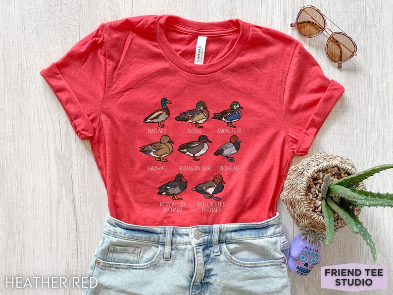 Peut inclure: Un t-shirt &agrave; manches courtes rouge chin&eacute; avec une impression graphique de neuf esp&egrave;ces de canards diff&eacute;rentes, accompagn&eacute;es de leurs noms respectifs : Mallard, Wood, Baikal Teal, Gadwall, Eurasian Teal, Redhead, Rosy Billed Pochard et Red Crested Pochard.